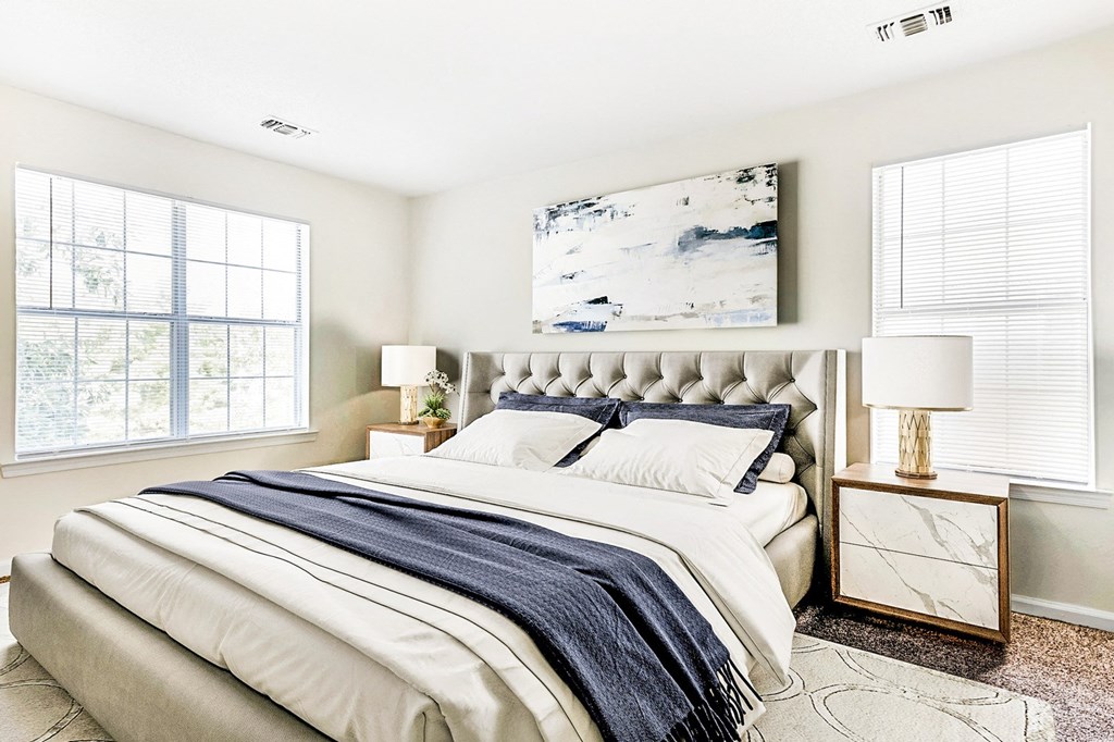 Staged bedroom with queen sized bed, art above bed, two nightstands, and large windows in the background.