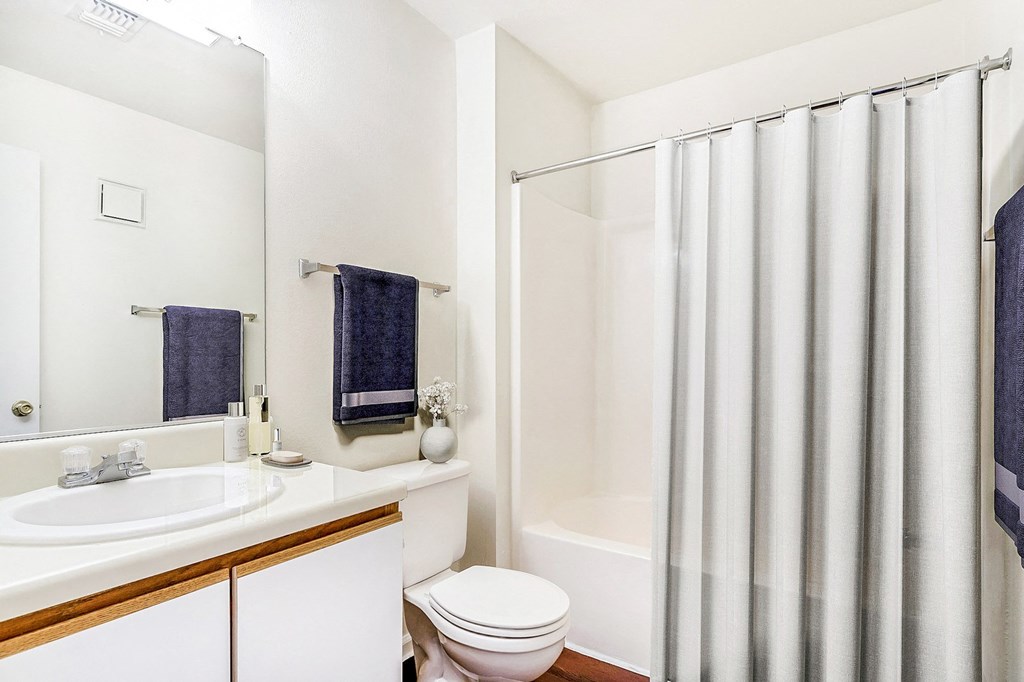 Staged bathroom with tiled bathtub shower combo. White counter tops, brown cabinets with a white faces, and wood style flooring. Blue towels hang on towel racks and lotions decorate the countertops.