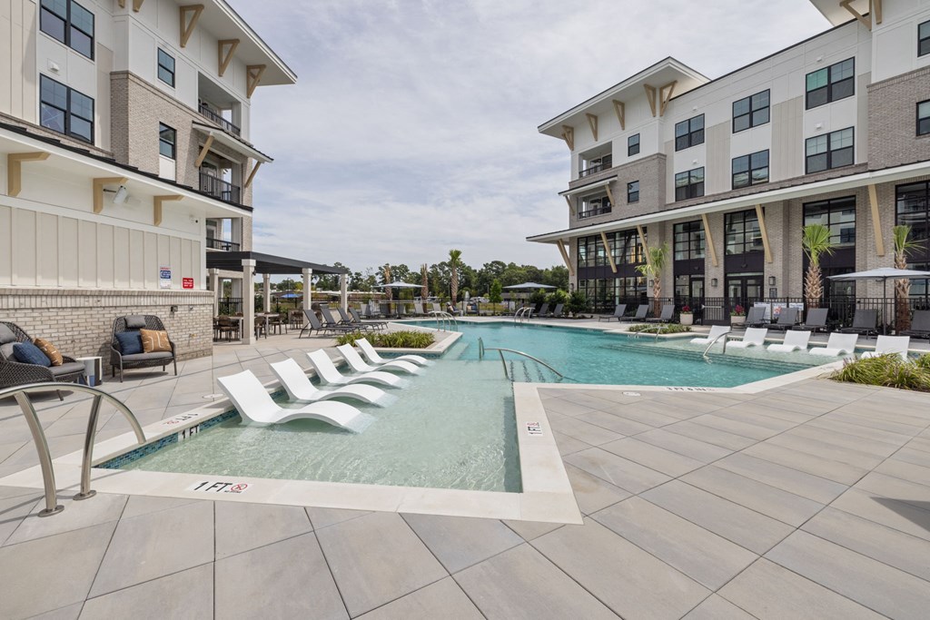 A large swimming pool surrounded by lounge chairs in front of apartment buildings.