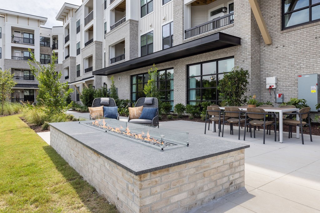 A patio with a fire pit and chairs is surrounded by a grassy area and apartment buildings.