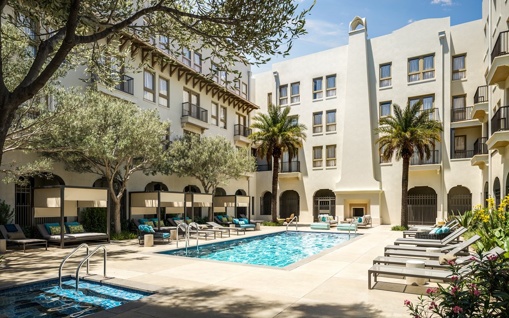 A pool area with sun loungers and a tree in front of a white building with balconies.