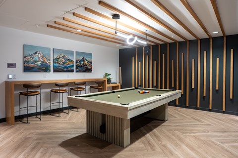 A pool table is in the middle of a room with a bar and pictures on the wall.