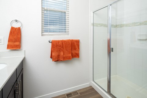A bathroom with a white sink, a window with blinds, a towel rack with two orange towels, and a glass shower door.