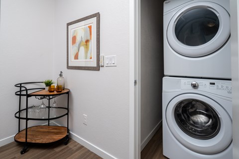 A washing machine is placed next to a wall with a picture frame and a cart with a bottle and a glass.