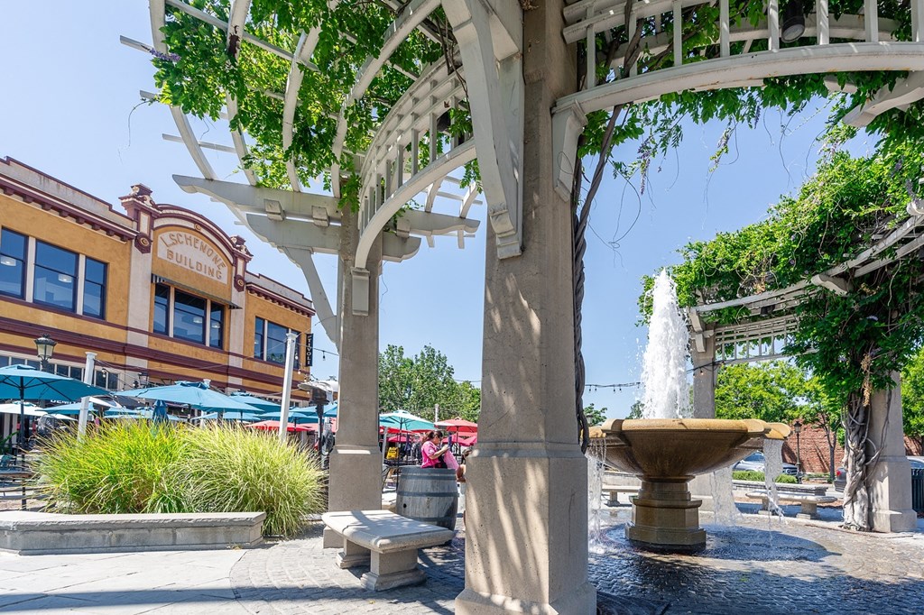 a fountain in the middle of a plaza with a pergola and a building in the