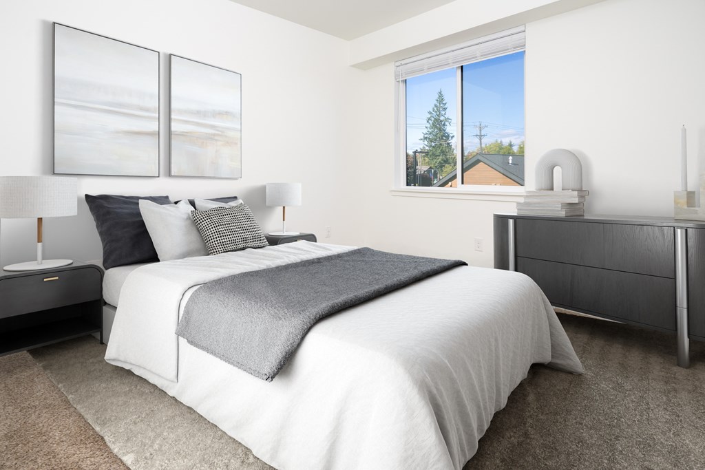 A bedroom with a large bed, a nightstand, and a window with a view of a house.