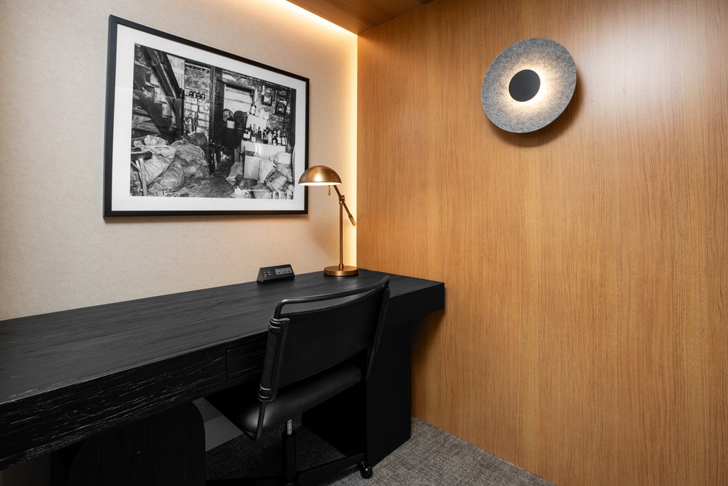 A black chair sits in front of a desk with a lamp and a picture on the wall.