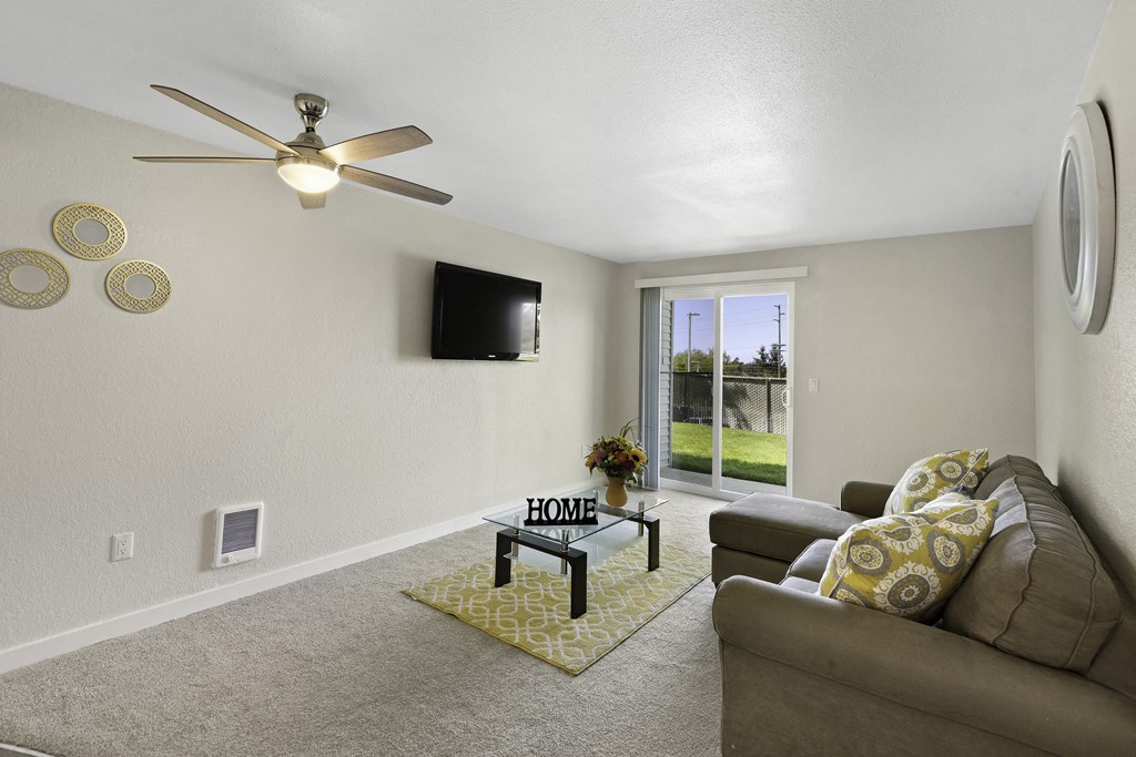 A Furnished Living Room with Carpeting, Sliding Glass Doors Leading to Outdoor Area, and Overhead Ceiling Fan with Lighting at Campo Basso Apartment Homes, Lynnwood, WA