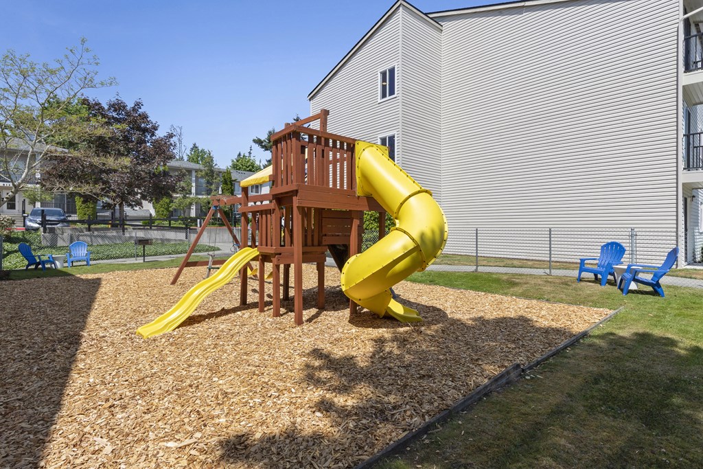 Property Playground with a yellow slide and a wooden deck with blue chairs in an outdoor area at Park Edmonds Apartment Homes, Edmonds, Washington