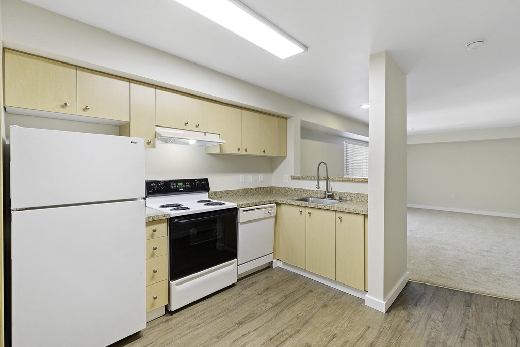 Kitchen with Plank Flooring that Includes a White Refrigerator with Separate Freezer Next to a Stove Top Oven, Wooden Cabinetry, Looking out into the Living Room at Excalibur Apartment H, Bellevue, WA