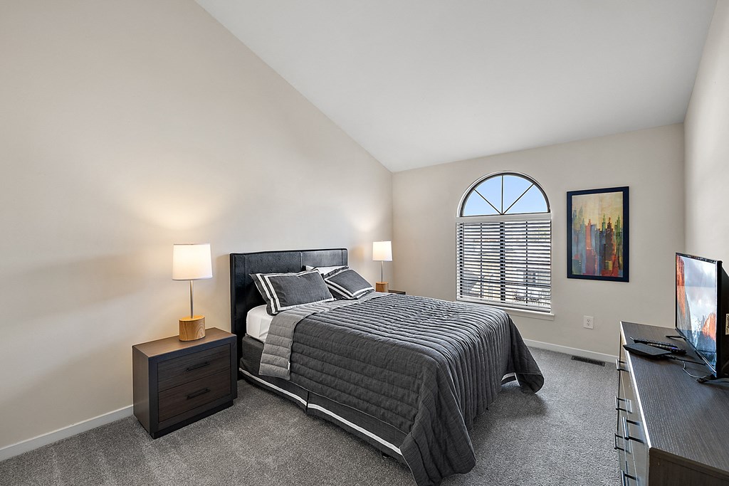 A bedroom with a large bed, a nightstand with a lamp, a window with blinds, and a painting on the wall at MainCentre, Northville