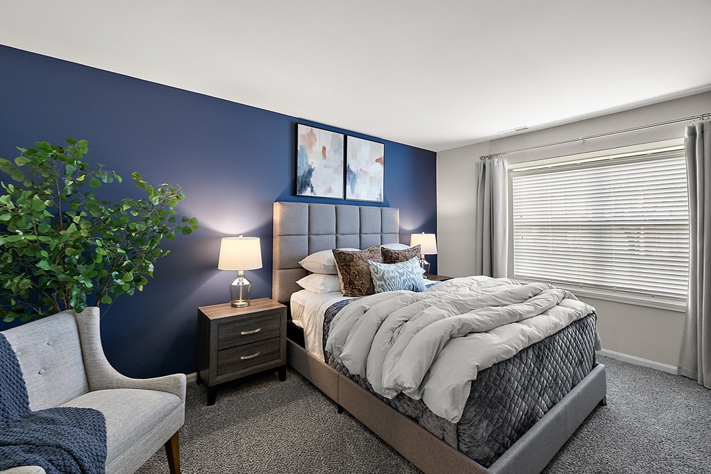 A bedroom with a bed, nightstand, chair, and a plant.