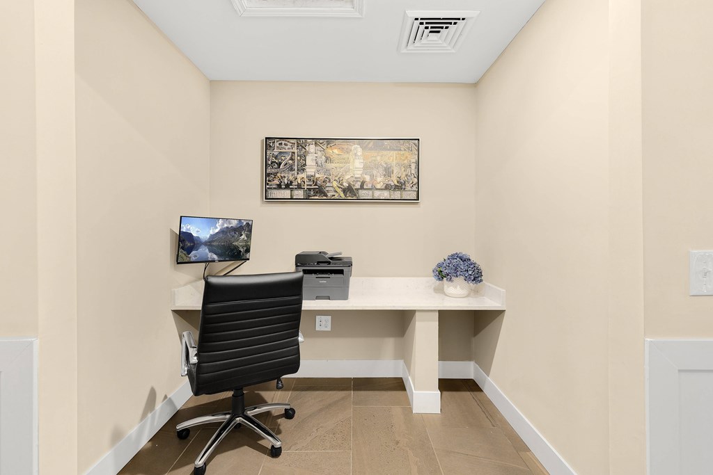 A black office chair is in front of a desk with a computer monitor and a framed picture on the wall.