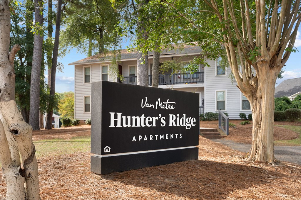 A sign for Hunter's Ridge Apartments is in front of a tree and a house.