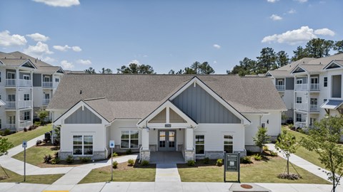 A large white building with a grey roof is surrounded by smaller buildings and trees.