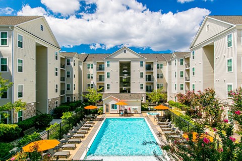 A large swimming pool surrounded by lounge chairs and umbrellas in front of apartment buildings.