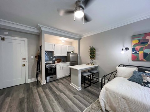 A bedroom with a bed, a table, and a kitchen area in the background.