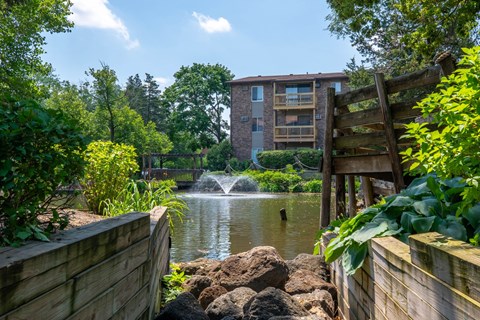 A wooden fence borders a pond with a waterfall and a building in the background.