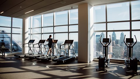 A person is exercising on a treadmill in a brightly lit gym with a view of the city.