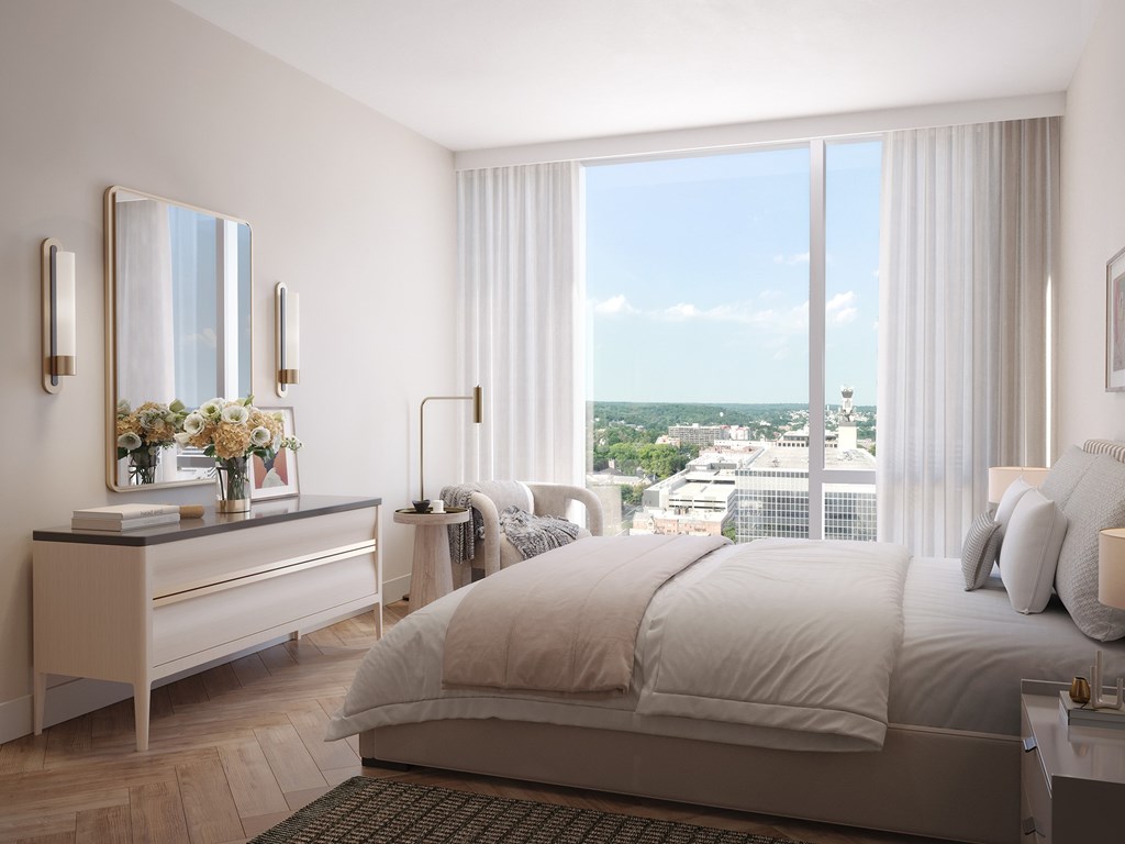 A bedroom with a large bed, a dresser with a mirror, and a view of the city outside the window.