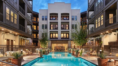 A large swimming pool is surrounded by potted plants and lounge chairs in the middle of a courtyard.
