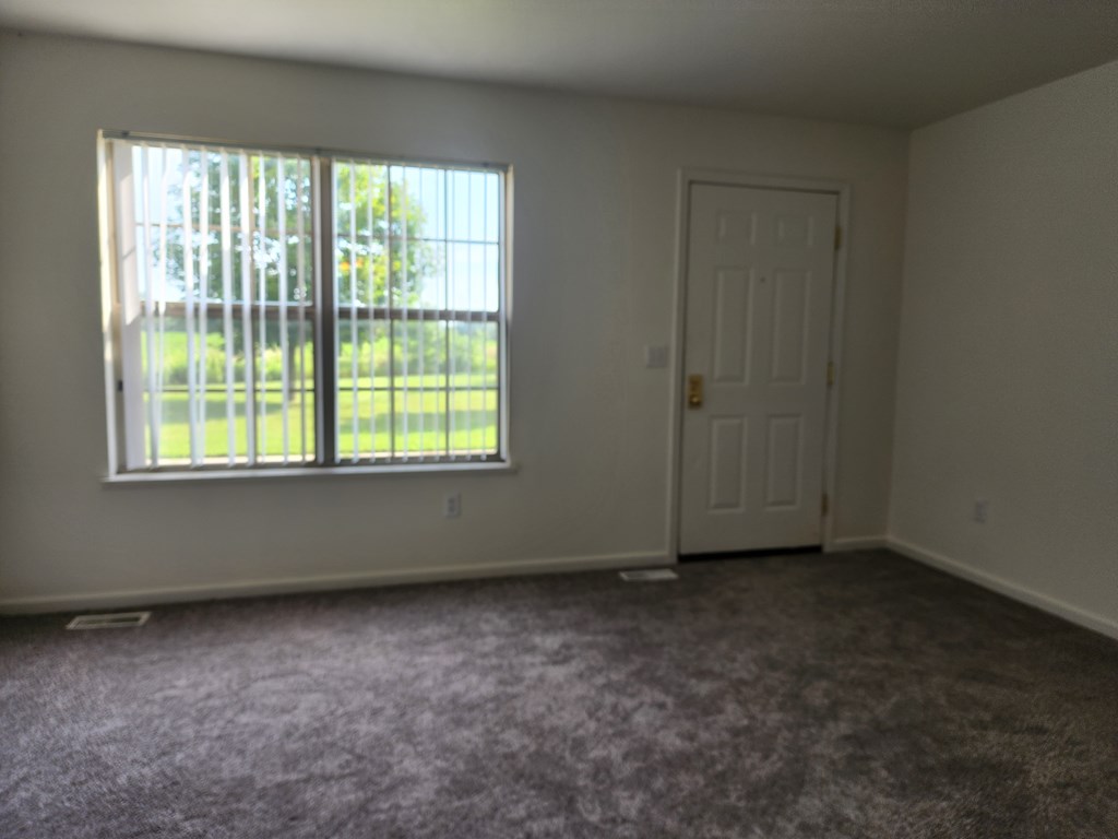 A room with a carpeted floor, a window with a view of a fence and trees, and a white door.