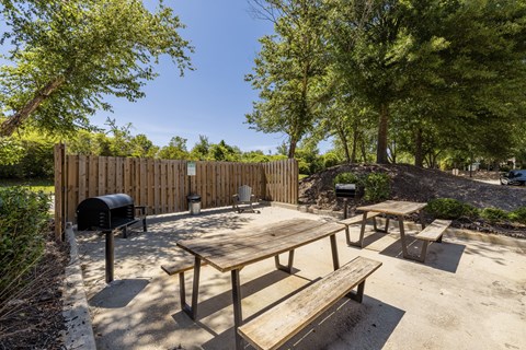 A wooden picnic table with benches is set in a sandy area with trees and a wooden fence in the background.
