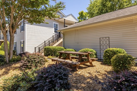 A wooden picnic table is in the middle of a garden with a house in the background.