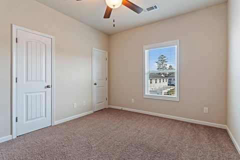 A room with a carpet floor, a ceiling fan, and a window with a view of a building and trees.