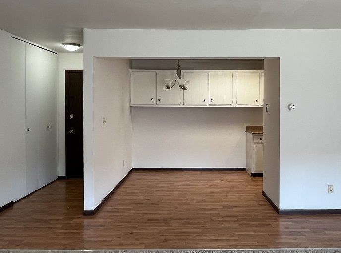 A room with white walls and wooden floors has a black door on the left and a white cabinet on the right.