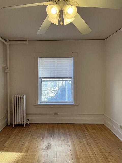 A room with a ceiling fan, a window with blinds, and a radiator on the floor.