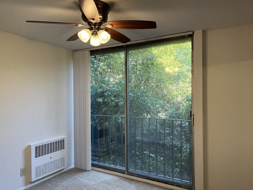 A room with a ceiling fan, a window with a view of trees, and a radiator on the wall.