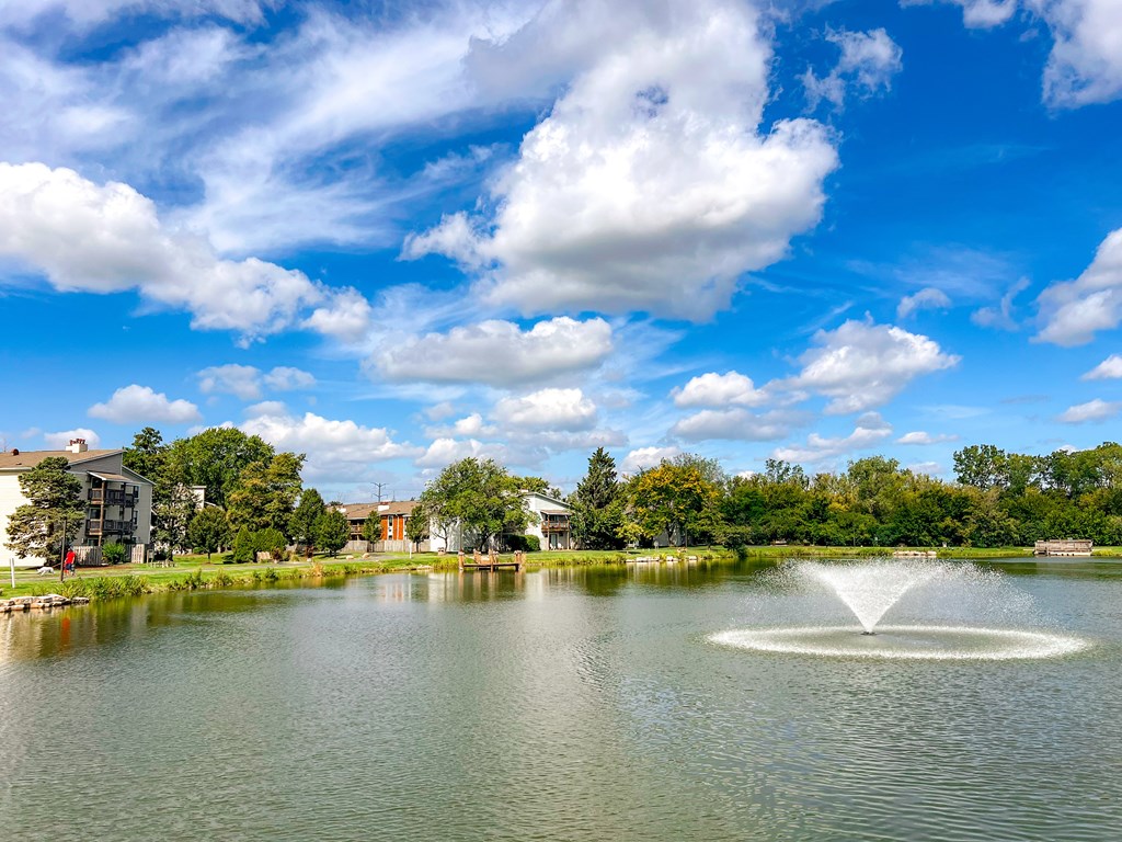A fountain in the middle of a lake with buildings and trees in the background.
