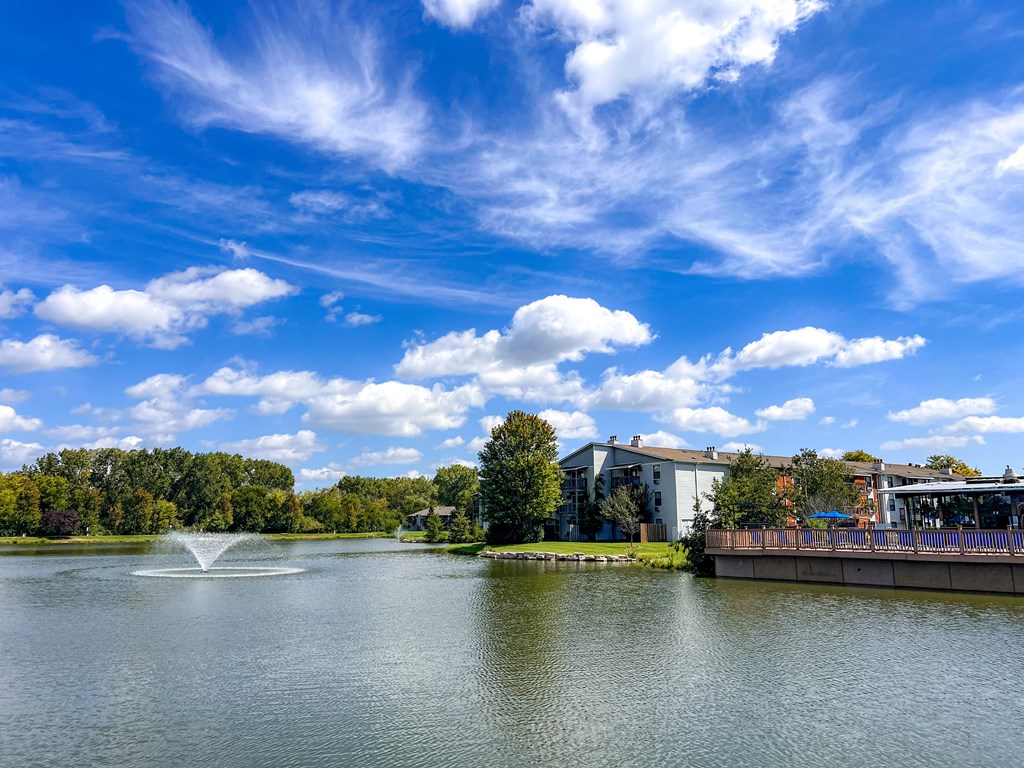 A serene lake with a fountain in the middle, surrounded by trees and buildings under a clear blue sky.