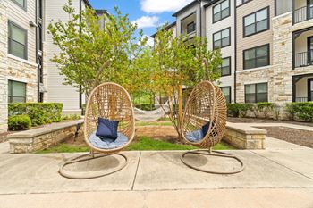 Two wicker chairs with blue cushions are placed on a concrete surface in front of apartment buildings.