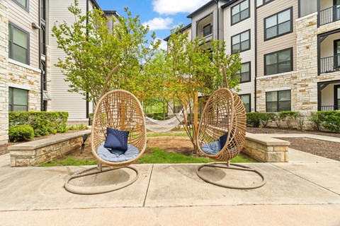 Two wicker chairs with blue cushions are placed on a concrete surface in front of apartment buildings.