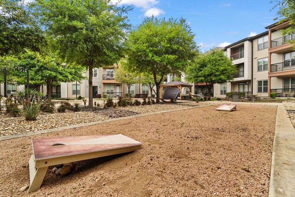 A playground with a seesaw in the middle of a gravel area with apartment buildings in the background.
