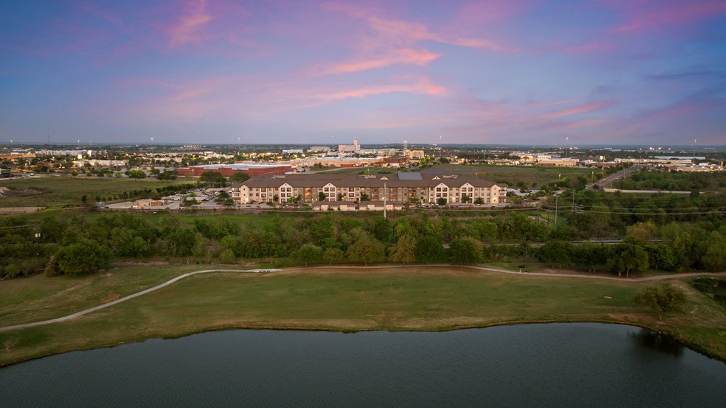 A large building complex is surrounded by a grassy field and a body of water under a pink and blue sky.
