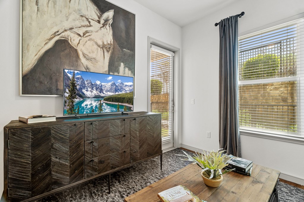 A room with a wooden cabinet, a painting of a giraffe, a TV displaying a mountain landscape, a window with a grey curtain, and a wooden table with a plant and a phone on it.