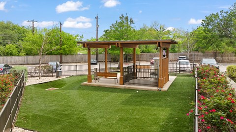 A wooden pavilion with a bench is surrounded by a green lawn and a fence.