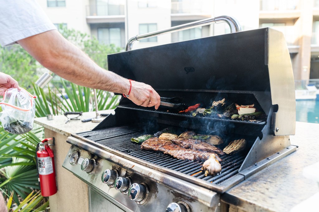 A man grilling meat on a barbecue with a red fire extinguisher nearby.