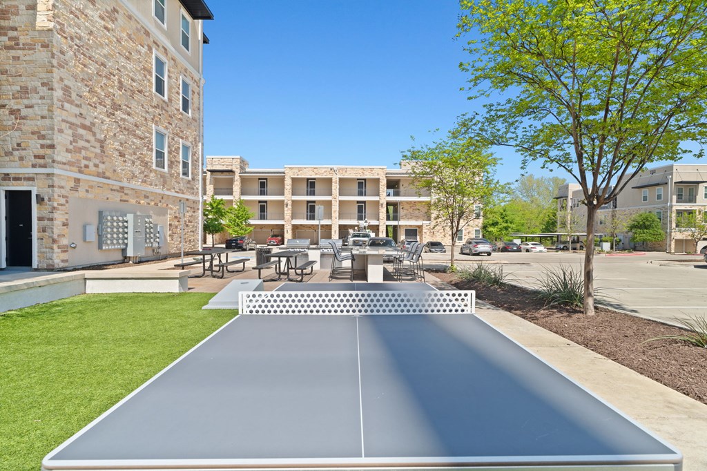 A shiny, metallic ping pong table is in the foreground of a sunny courtyard.