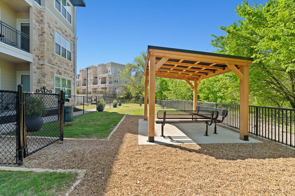 A wooden pergola with benches is situated in a gravel area.