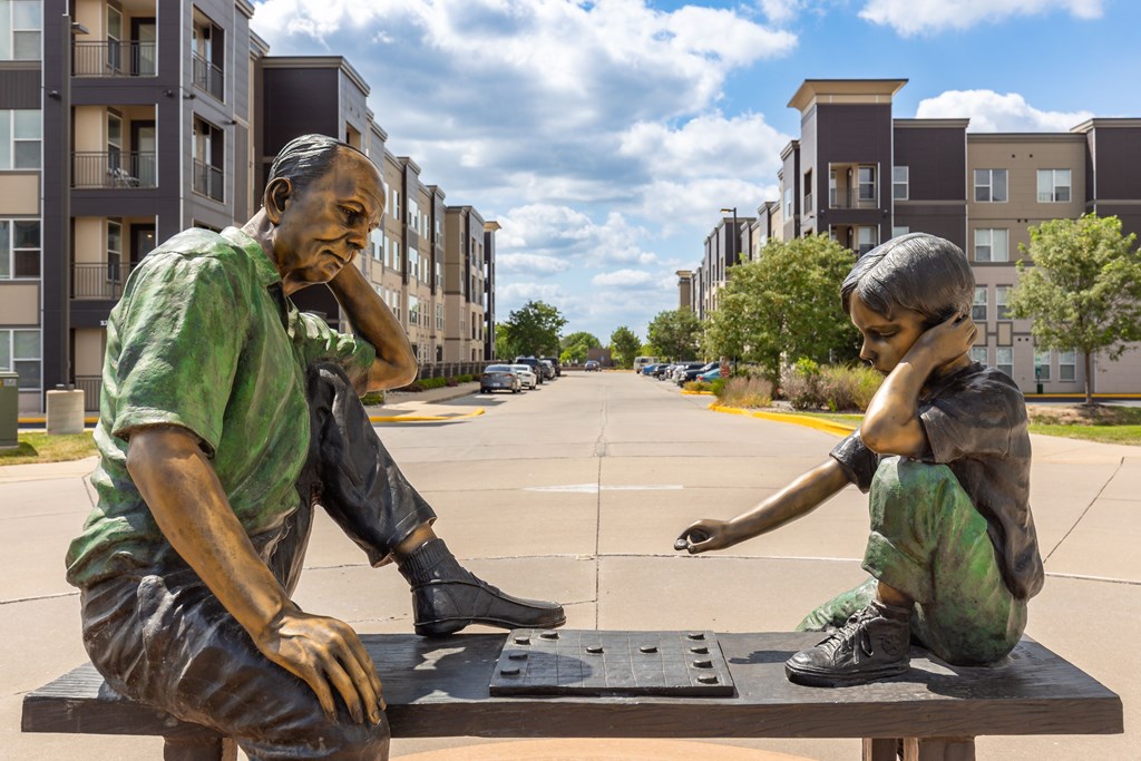A statue of two people sitting on a bench in front of apartment buildings at Alinea Town and Country Luxury Apartments in Des Peres MO 63131..