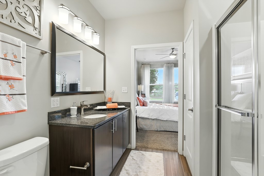 A bathroom with a sink, mirror, and a bedroom in the background at Alinea Town and Country Luxury Apartments in Des Peres MO 63131..