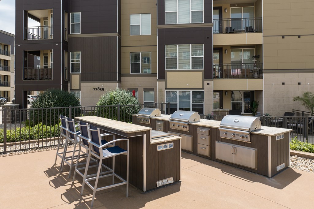 A patio with a grill and chairs is in front of apartment buildings at Alinea Town and Country Luxury Apartments in Des Peres MO 63131..