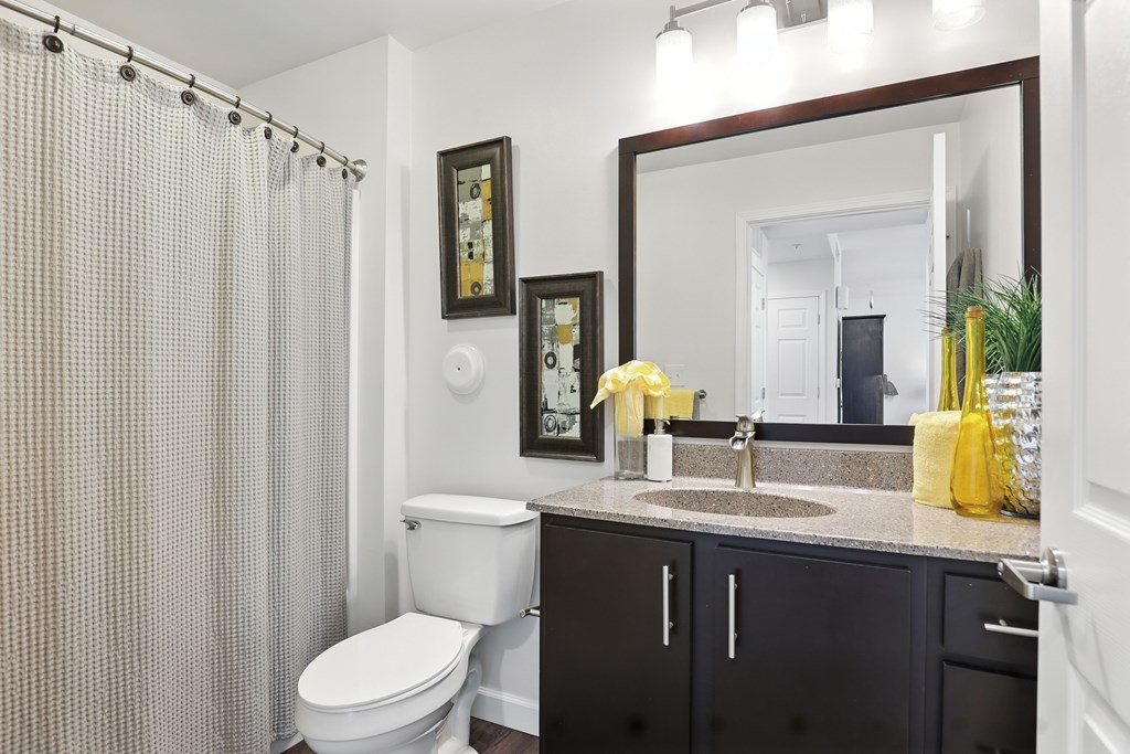 A bathroom with a white toilet, a sink with a yellow vase, and a shower curtain at Bramblett Hills Luxury Apartments in O Fallon MO 63366