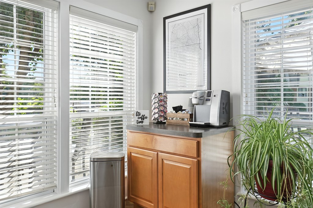 A room with a window covered in blinds, a plant, a framed map, and a microwave on a counter.