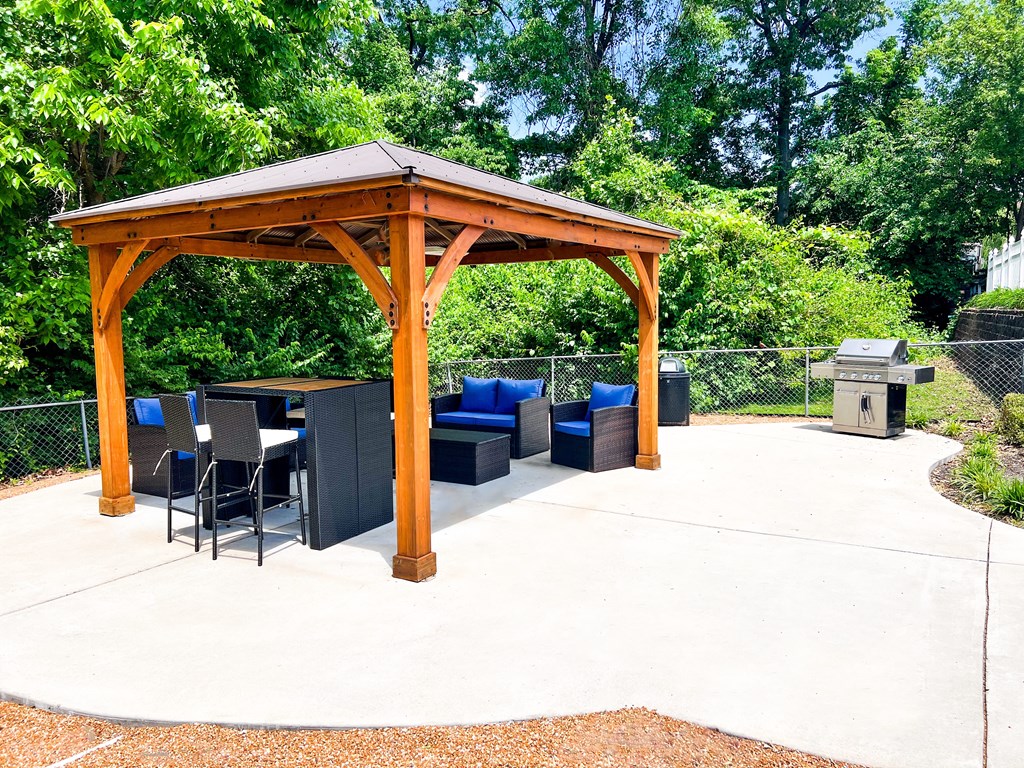 A wooden gazebo with a table and chairs is surrounded by trees.