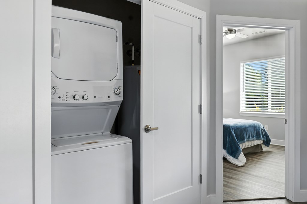 A white washing machine is in a small room with a bed in the background.